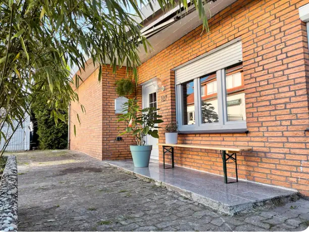 Fassade Senne Bungalow Backsteinfassade eines Hauses mit weißem Eingang, Fensterläden und Topfpflanze auf gepflastertem Weg.