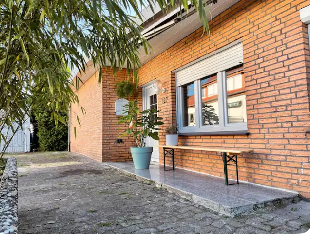 Fassade Senne Bungalow Backsteinfassade eines Hauses mit weißem Eingang, Fensterläden und Topfpflanze auf gepflastertem Weg.