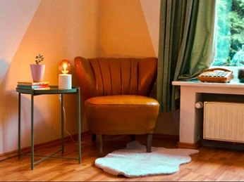 Wohnzimmer Senne Bungalow Ein lederner Sessel neben einem kleinen Tisch mit Lampe und Pflanze, vor einem Fenster mit Vorhang.