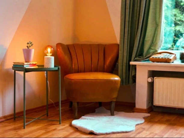 Wohnzimmer Senne Bungalow Ein lederner Sessel neben einem kleinen Tisch mit Lampe und Pflanze, vor einem Fenster mit Vorhang.