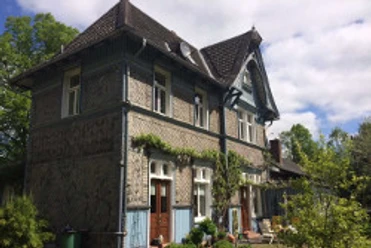 altstdtbhf-1.jpg Historischer Bahnhof in Fachwerkstil mit begrünten Fassaden, umgeben von Bäumen und blauen Himmel.