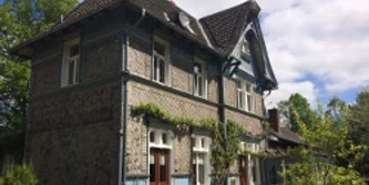 altstdtbhf-1.jpg Historischer Bahnhof in Fachwerkstil mit begrünten Fassaden, umgeben von Bäumen und blauen Himmel.