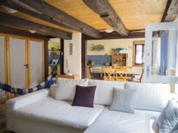 wohnraum1.jpg Wohnzimmer mit weißem Sofa, Holzbalkendecke und farbiger Küche im Hintergrund. Gemütliche Atmosphäre.