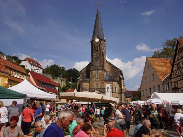 Menschen versammeln sich auf einem belebten Wochenmarkt vor einer Kirche mit spitzem Kirchturm.