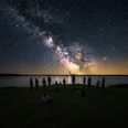 gemini-bz.png Gruppe von Menschen betrachtet am Seeufer den klaren Sternenhimmel mit leuchtender Milchstraße.