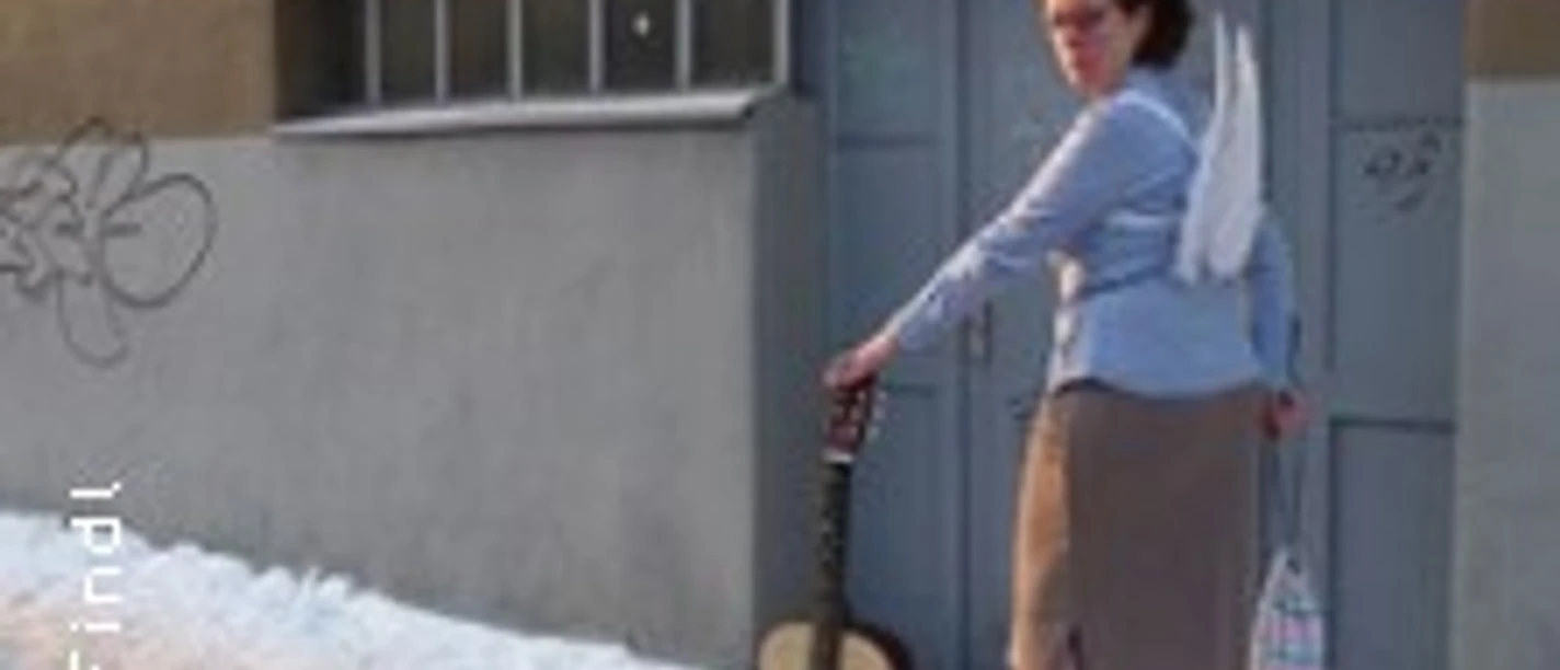Eine Frau mit einem Engel-Kostüm aus Hemd, Rock und Flügeln hält eine Gitarre vor einer Garagenwand.