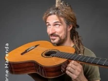 Ein Mann mit Dreadlocks hält eine Gitarre und blickt lächelnd nach oben, vor grauem Hintergrund.
