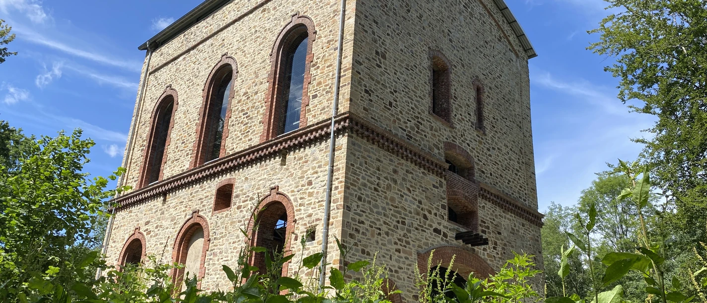 Stueveschacht_Bildrechte_Cornelia Saure_Stadt_OS.JPG Historic brick building with arched windows, surrounded by lush greenery and blue skies.
