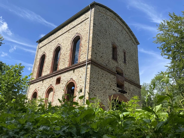 Stueveschacht_Bildrechte_Cornelia Saure_Stadt_OS.JPG Historisches Backsteingebäude mit rundbogigen Fenstern, umgeben von üppigem Grün und blauem Himmel.