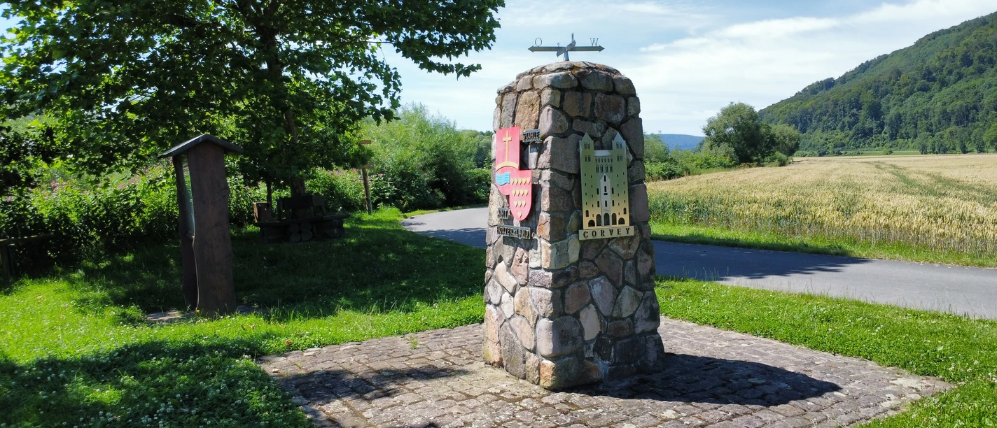 oestlichster_winkel_nrw_stahle_struck.jpg Steinerner Infopunkt am Rand eines Radweges mit angebrachtem Wappen der Ortschaft und Silhouette des Corveyer Westwerkes