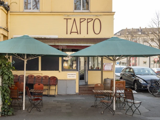 Tappo Außenaufnahme des Restaurants Tappo in Köln mit grünen Sonnenschirmen und leeren Sitzplätzen.