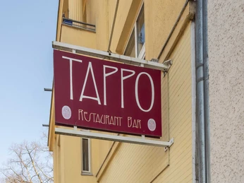Tappo Außenansicht des Tappo Restaurants mit rotem Schild, gelber Fassade und angrenzendem Gehweg.Exterior view of the Tappo restaurant with red sign, yellow facade and adjacent sidewalk.