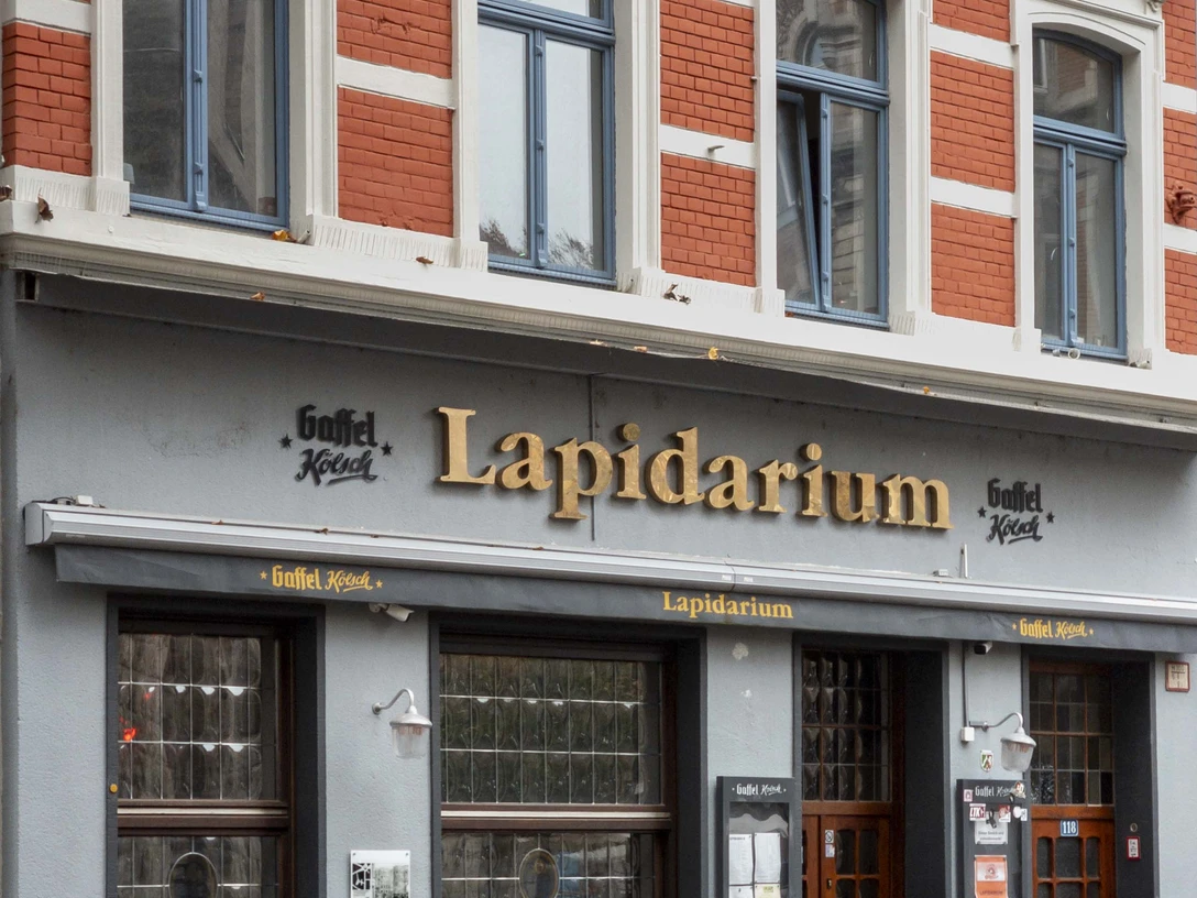lapidarium-KoelnTourismus-Seelbach-24.jpg Ein Eingang des Lapidarium in Köln mit roten Ziegeln und blauen Fensterrahmen.Facade of a historic pub named "Lapidarium" with outdoor seating and brick architecture.