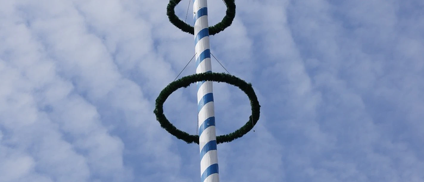 Blau-weiß gestreifter Maibaum ragt in den bewölkten Himmel, geschmückt mit grünen Ringen.