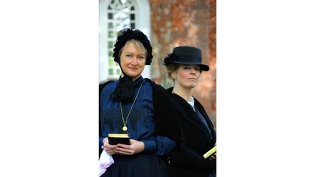 Pastorengattinnen Two women dressed in Victorian style in front of a brick-red wall.