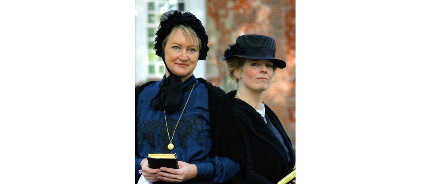 Pastorengattinnen Two women dressed in Victorian style in front of a brick-red wall.