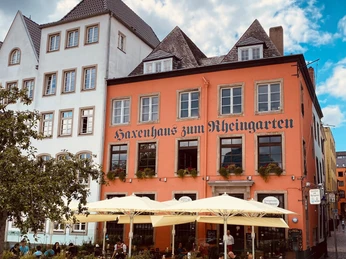 Haxenhaus Personen sitzen unter Sonnenschirmen vor einem historischen Fachwerkhaus mit der Aufschrift "Haxenhaus zum Rheingarten".People sit under parasols in front of a historic half-timbered house with the sign "Haxenhaus zum Rheingarten".