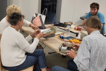 Personen arbeiten an einem Tisch mit alten Elektrogeräten und Werkzeugen in einem Workshop.