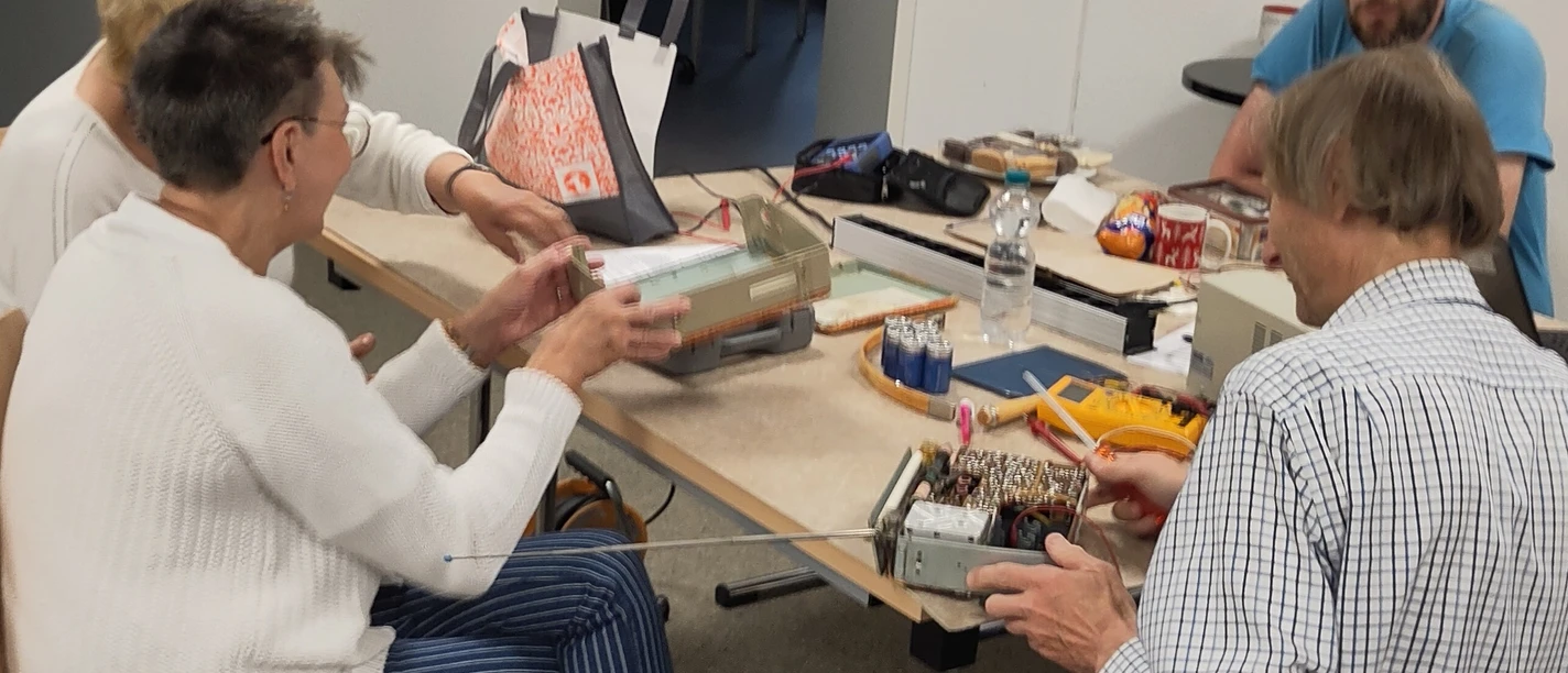 Personen arbeiten an einem Tisch mit alten Elektrogeräten und Werkzeugen in einem Workshop.