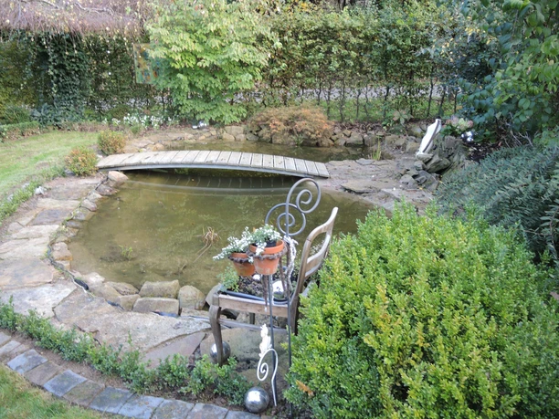Kleiner Teich mit Holzbrücke, umgeben von Pflanzen und einem Gartenstuhl mit Blumentopf.