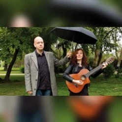 Zwei Personen im Park, eine hält eine Gitarre, die andere einen Regenschirm über beide.