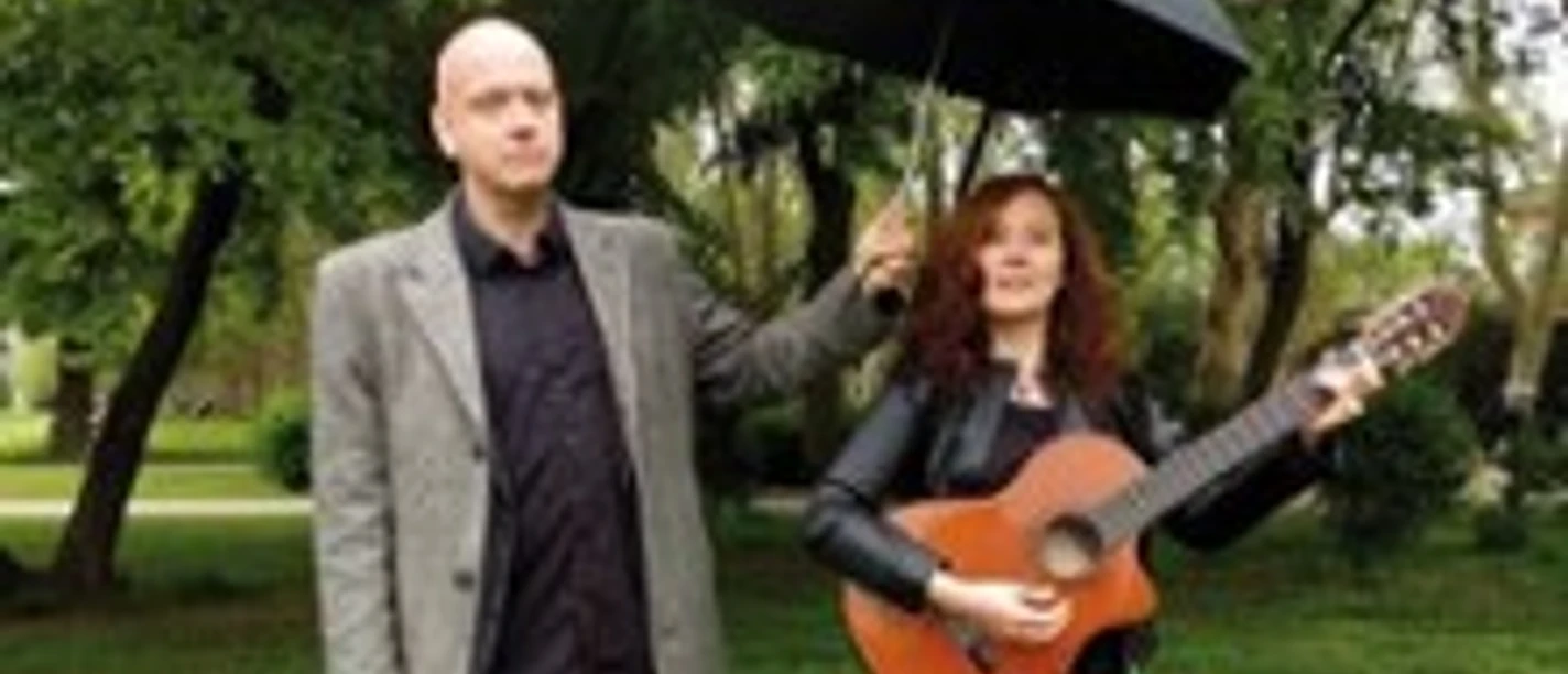 Zwei Personen im Park, eine hält eine Gitarre, die andere einen Regenschirm über beide.