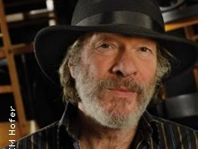 Mann mit Hut und Bart lächelt sanft in die Kamera, umgeben von einer gemütlichen, warmen Atmosphäre.