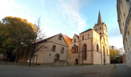 HF_Muenster1_Siwinski.jpg Historische Kirche mit gotischem Turm, umgeben von Herbstbäumen und Kopfsteinpflasterweg.