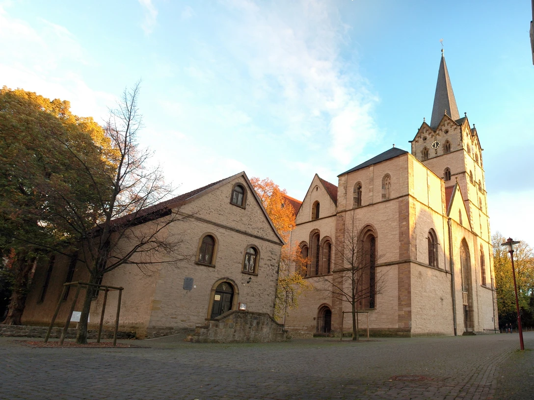 HF_Muenster1_Siwinski.jpg Historische Kirche mit gotischem Turm, umgeben von Herbstbäumen und Kopfsteinpflasterweg.
