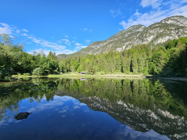 Schmölzer See Du siehst einen klaren See mit Wald, Bergen und einer kleinen Hütte.