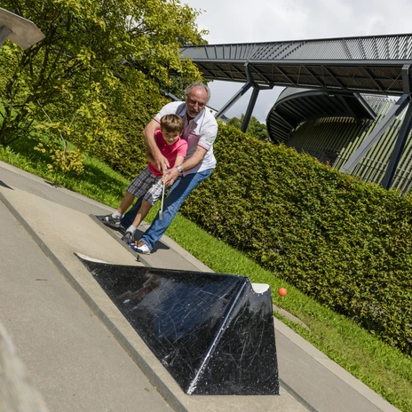 A man plays miniature golf with his grandchild, in the background the multifunctional hall "das O", leisure, zoo, family outing
