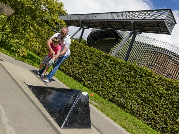 A man plays miniature golf with his grandchild, in the background the multifunctional hall "das O", leisure, zoo, family outing