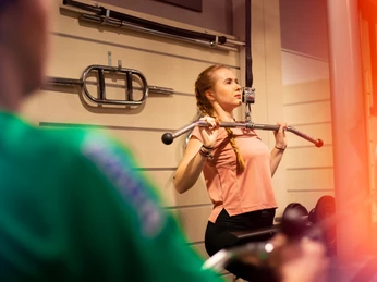 Eine Frau im Fitnessstudio trainiert intensiv an einem Latzuggerät, umgeben von Sportausrüstung.A woman in the gym is working out intensively on a lat pull-down machine, surrounded by sports equipment.