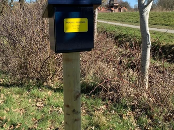 Stempelbox Foto Antje Geisler.jpg Das Bild zeigt eine blaue Stempelbox auf einem Holzpfosten in einer ländlichen Umgebung mit Bäumen und einer Windmühle im Hintergrund.