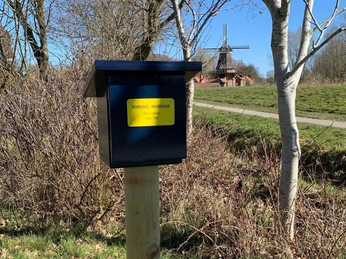Stempelbox Foto Antje Geisler.jpg Das Bild zeigt eine blaue Stempelbox auf einem Holzpfosten in einer ländlichen Umgebung mit Bäumen und einer Windmühle im Hintergrund.