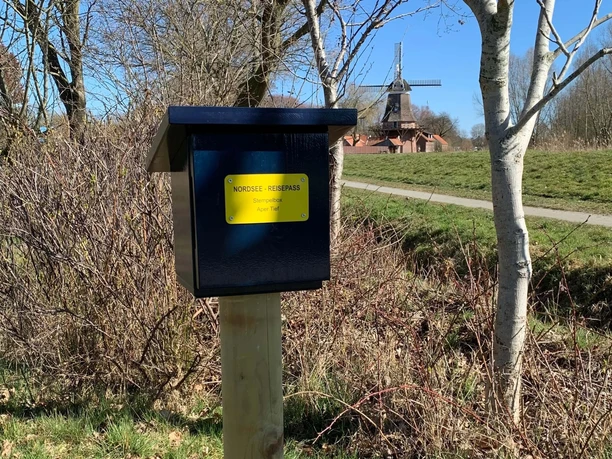 Stempelbox Foto Antje Geisler.jpg Das Bild zeigt eine blaue Stempelbox auf einem Holzpfosten in einer ländlichen Umgebung mit Bäumen und einer Windmühle im Hintergrund.