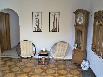 IMG-20250321-WA0012.jpg Zwei geflochtene Sessel und ein Glastisch mit Dekoration in einem Raum mit antiker Standuhr.