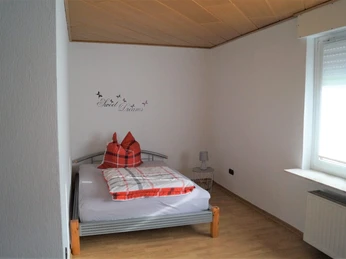IMG-20250321-WA0008.jpg Ein schlichtes Schlafzimmer mit Einzelbett, roten Kissen, Fenster mit Rolladen, Holzboden und kleiner Lampe.