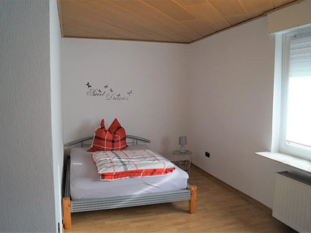 IMG-20250321-WA0008.jpg Ein schlichtes Schlafzimmer mit Einzelbett, roten Kissen, Fenster mit Rolladen, Holzboden und kleiner Lampe.