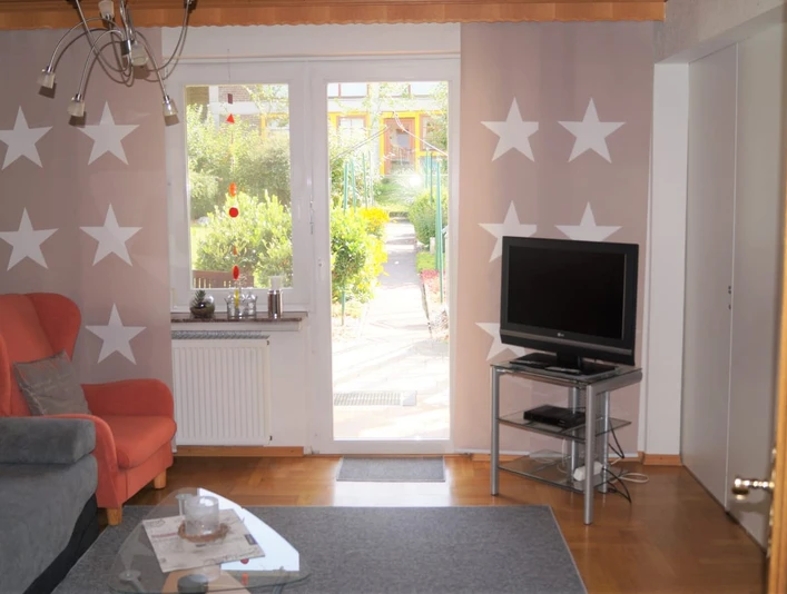 IMG-20250321-WA0010.jpg Ein Wohnzimmer mit einem grauen Sofa, einem roten Sessel und einem Fernseher vor einem großen Fenster.