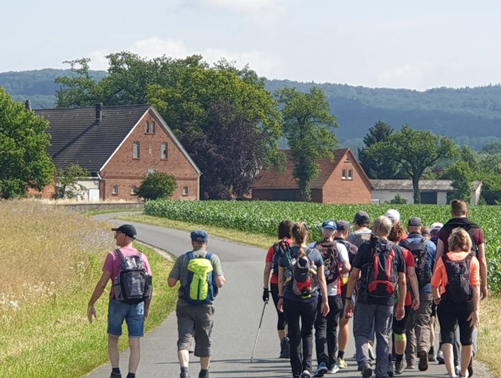 Wfg_Marsch_Wandergruppe.jpg Eine Gruppe von Wanderern mit Rucksäcken spaziert auf einer ländlichen Straße vorbei an Häusern.