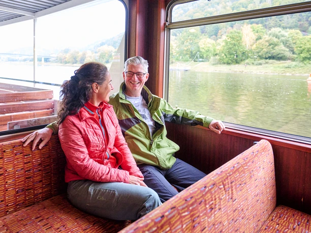 A man and a woman sit smiling on a boat bench, he wears a green jacket, she a red one, with a view of a river and green riverbank landscape.
