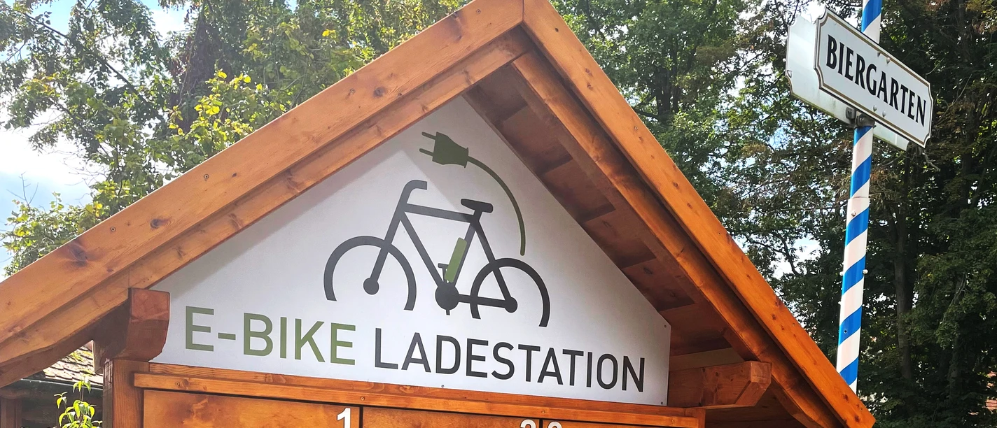 Holzhütte mit E-Bike-Ladestation, umgeben von Bäumen; nahe einem Biergarten gelegen.