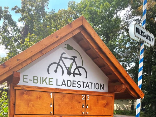Holzhütte mit E-Bike-Ladestation, umgeben von Bäumen; nahe einem Biergarten gelegen.