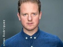 Ein Mann im blauen Hemd mit kurzen, hellen Haaren blickt ernst in die Kamera vor grauem Hintergrund.