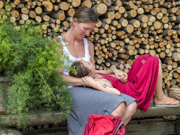 Een vrouw in een wit topje houdt een kind in een rode jurk op haar schoot, voor een stapel boomstammen, naast een groene struik.