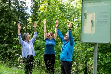 Atemwegslehrpfad Drei Menschen heben Arme im Wald. Du siehst eine Übungstafel daneben.