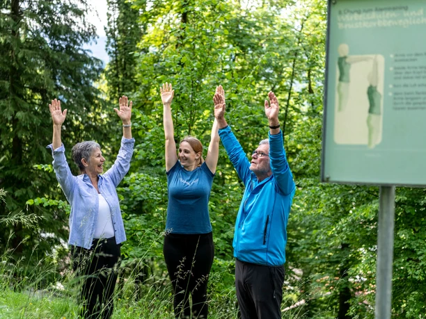 Atemwegslehrpfad Drei Menschen heben Arme im Wald. Du siehst eine Übungstafel daneben.