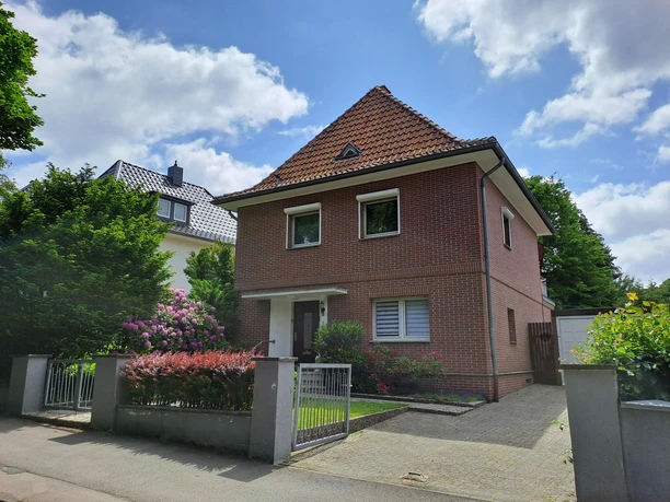 Ein gemütliches, zweigeschossiges Haus mit rotem Ziegeldach und einem gepflegten Garten. Umgeben von grünen Pflanzen und einer einladenden Atmosphäre.