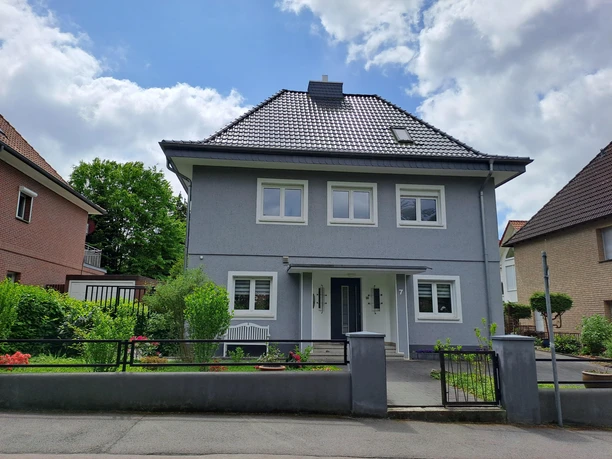 Ein modernes, graues Haus mit einem Satteldach und weißen Fenstern. Vor dem Haus gibt es einen kleinen Garten und eine gepflegte Einfahrt.